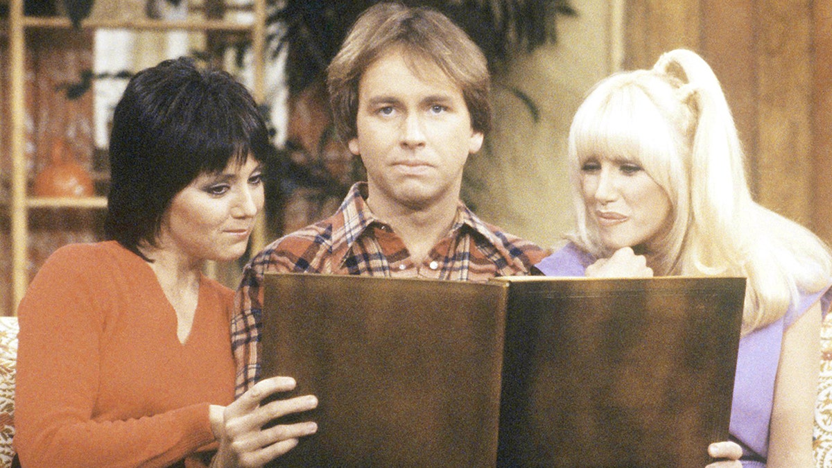 Three's Company cast Joyce DeWitt (Janet), John Ritter (Jack) and Suzanne Sommers (Chrissy) are seated on the couch, with the girls looking at a brown book and Ritter looking directly at the camera