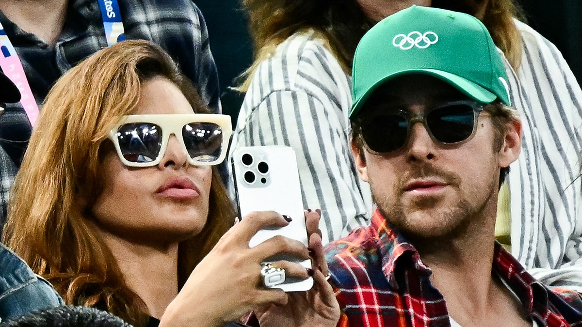 Eva Mendes holds up a white iPhone while wearing cream sunglasses sitting next to husband Ryan Gosling in a plaid shirt and green hat at the US Open