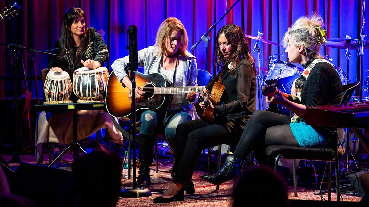 Meena Makhijani sits on stage with The Bangles members Vicki Peterson and Susana Hoffs, both holding guitars and Annette Zalinskis facing inward also holding a guitar