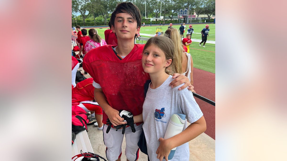 Benjamin Brady in a red shirt at a football game/practice stands tall next to his sister Viivan in a white t-shirt