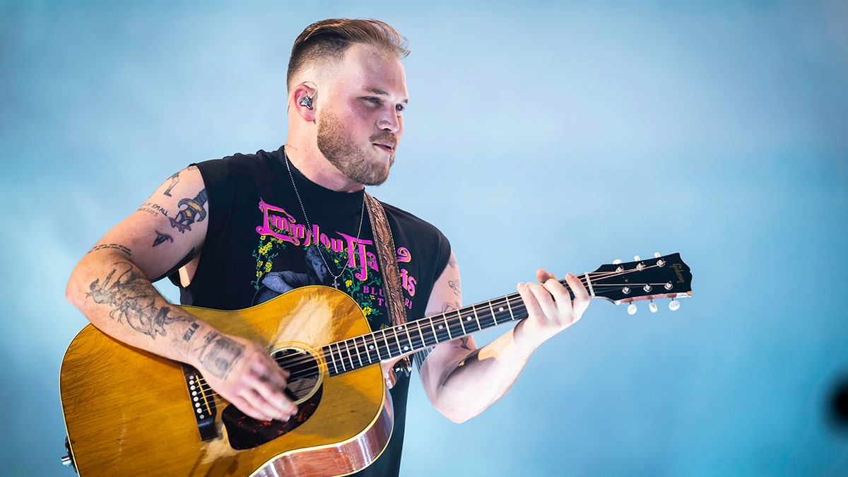 Zach Bryan in a cut-off black tank top strumming his guitar on stage