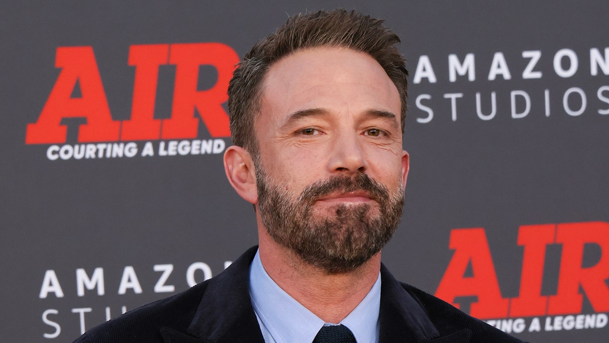 Ben Affleck soft smiles on the carpet wearing a black suit and blue shirt