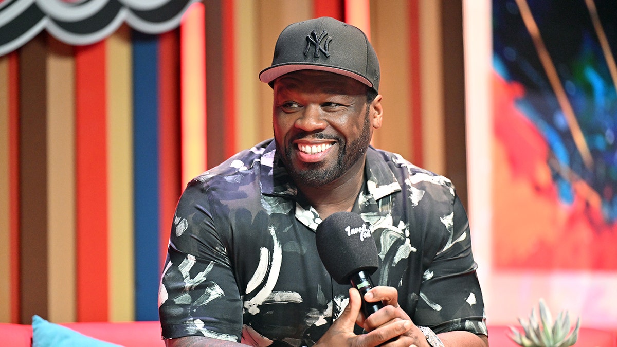 50 cent in a black New York Yankees baseball cap and patterned shirt smiles holding a microphone on stage