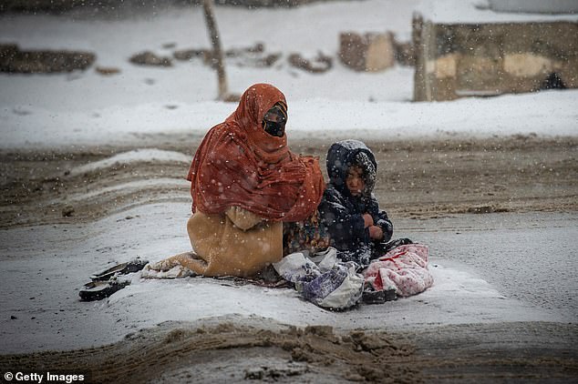 An Afghan woman begs for money from passing cars in the snow, with a child huddled beside her, on the Kabul road south to Pul-e Alam, Afghanistan, on January 17, 2022