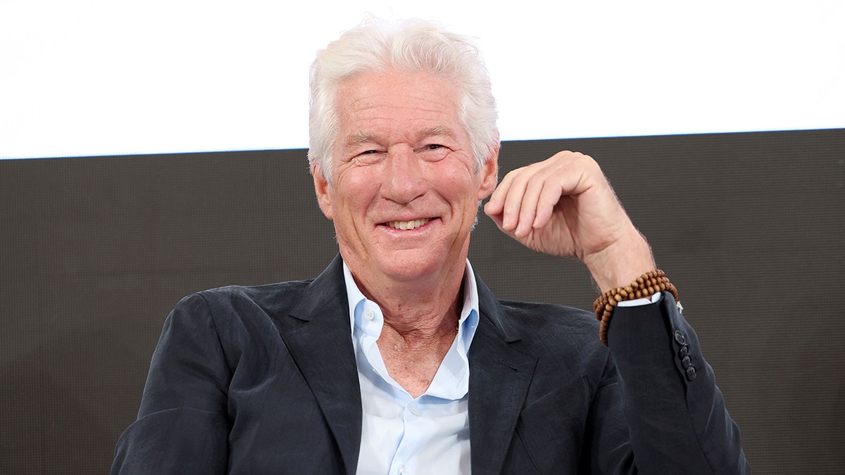 Richard Gere in a light blue shirt and black suit smiles as he sits on stage