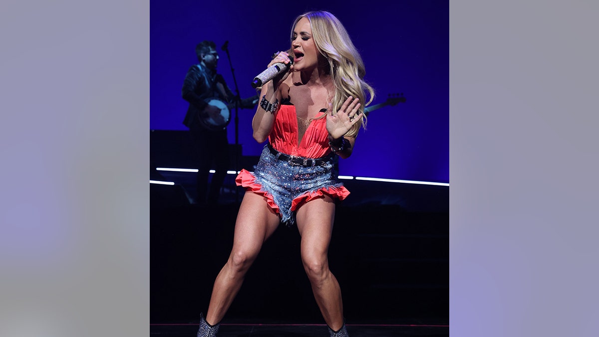 Carrie Underwood in a red bustier top and jean shorts with red trim sings passionately into the microphone