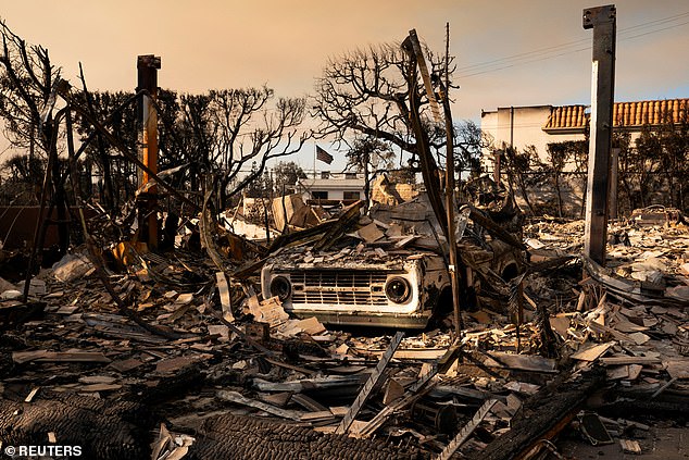 A partially burnt US flag flies behind the remains of a vehicle and home destroyed by the Palisades Fire