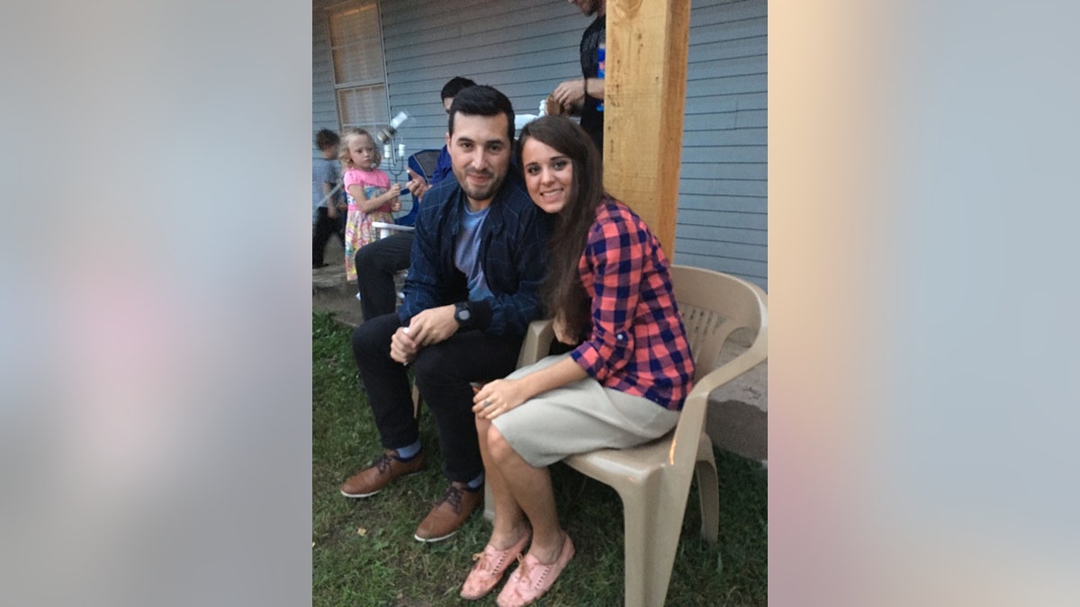 Jinger Duggar Vuolo in a blue and pink plaid shirt sitting on a beige chair and leaning towards her husband Jeremy Vuolo in a blue suit.